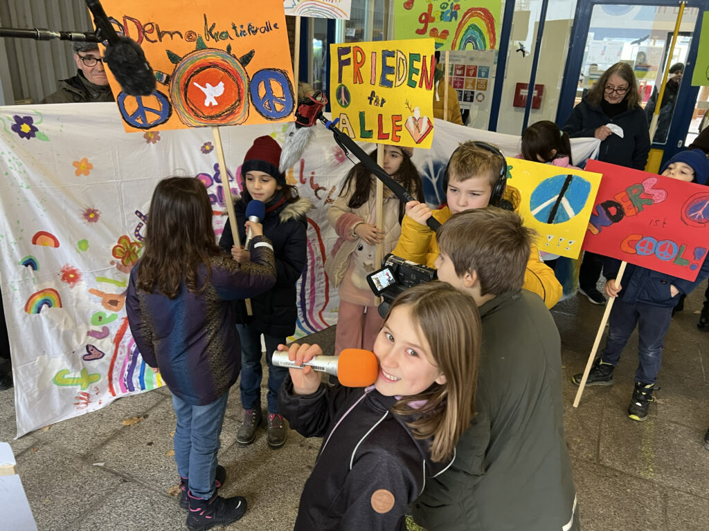 Grundschulkinder drehen einen Kurzfilm: Einige halten bunte Demonstrationsschilder, andere haben Kameras und Mikrofone in den Händen. Ein Mädchen mit Mikrofon vor dem Mund lächelt in die Kamera.