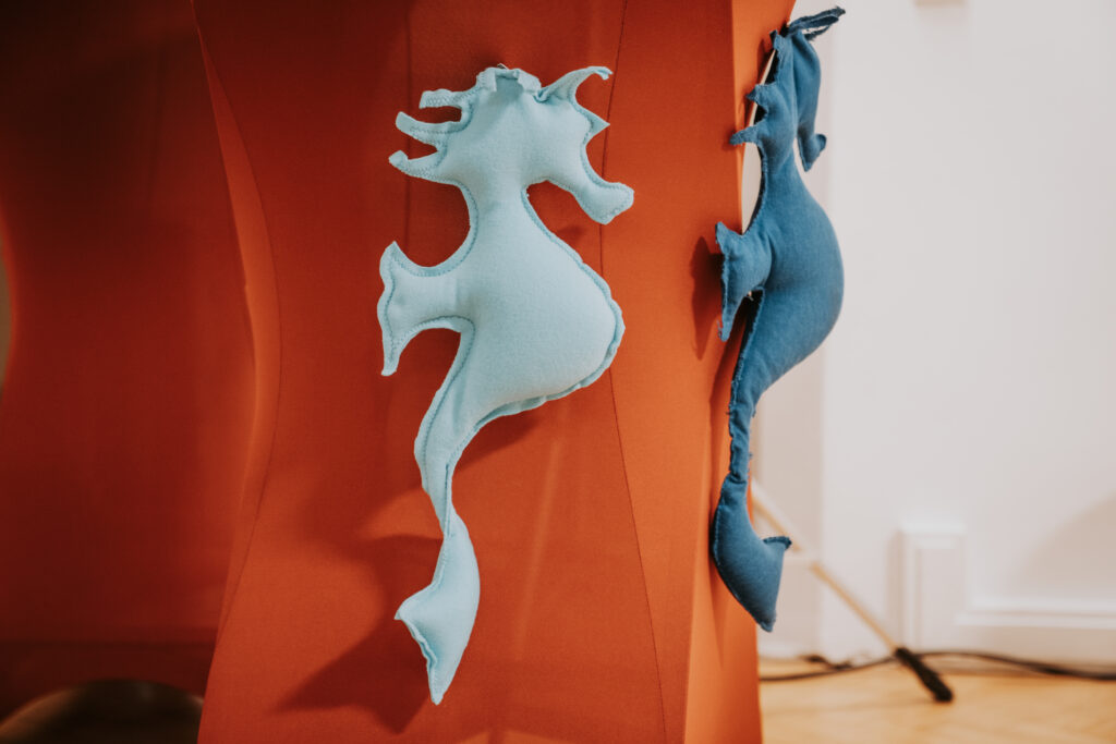 Zwei aus hellblauem und dunkelblauem Stoff genähte Seepferdchen hängen auf orangefarbenem Grund