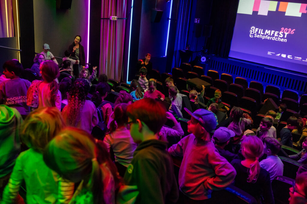 Kinder in einem Kinosaal, buntes Licht, auf der Leinwand steht: Filmfest Sehpferdchen 2026